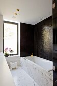 Bathtub with marble surround against wall with dark mosaic tiles in modern bathroom