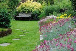 Verschiedene, blühende Blumen in sommerlichem Garten (im Voredergrund gewöhnliche Katzenpfötchen, hinter der Bank Spiere), seitlich Trittplatten als Weg in gemähter Rasenfläche