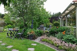 Gartenplatz mit Holztisch und -stühlen, seitlich angelegte Beete mit blühenden Blumen vor Anbau