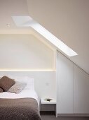 Knitted cushions and blankets in natural shades on bed in purist attic room with fitted wardrobe below sloping ceiling