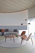 Klassikerstühle und Coffeetable aus Holz auf rundem Teppich vor massgefertigtem Sofa in Hellgrau, an gebogener Lounge Wand, seitlich Retro Stehleuchte