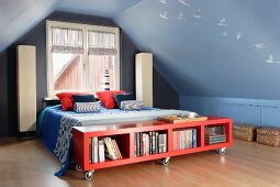 Rotes Lowboard auf Rollen vor Doppelbett, Stehleuchten an Giebelwand, in mittelblau getöntem Dachzimmer