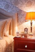 Nachttischleuchte mit gelbem Lampenschirm auf Nachtkästchen aus Mahagoniholz, neben teilweise sichtbarem Bett mit Kopfteil, an Wand helle Seidentapete mit Blumenmuster