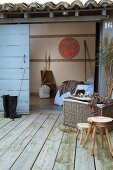 Wooden stools and wicker trunk on wooden terrace in front of sliding door leading to bedroom