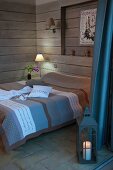Doppelbett mit gestreifter Tagesdecke in Brauntönen und Tischleuchte in Zimmerecke mit Holzverschalung, im Vordergrund Bodenlaterne