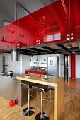 Wooden breakfast bar adjoining stainless steel island counter and bar stools below gallery with red glass balustrade in loft apartment