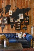 Vintage Ledercouch in Blau vor Holzwand mit aufgemalter Weltkarte, kleine Hängeschränke zwischen Bildern, im Vordergrund rustikale Gefässe auf Couchtisch
