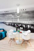 Tulip table, Eames Plastic Armchairs and blue sofa in front of black and white mural wallpaper depicting long wooden pier in the ocean