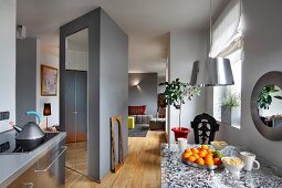 Grey kitchen counter with stainless steel fronts and dining table with floral pattern sandwiched in glass top; sculptural cupboard used as partition separating living area in background
