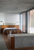 Concrete bathtub and double bed with brown bedspread in bedroom with concrete ceiling