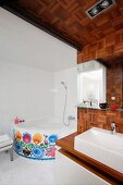 Modernes Bad, teilweise mit Mosaik Holzplatten an Wand und Decke ausgelegt, eingebauter Waschtisch, im Hintergrund Eckbadewanne mit dekorativem Blumenmuster an Frontseite
