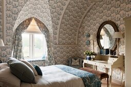 Oval mirror above antique desk with drawers on top and toile-de-jouy wallpaper and curtains in attic bedroom in historical manor house