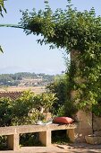Blick von Terrasse über schlichte Steinbank mit Rankbogen in sommerliche mediterrane Landschaft