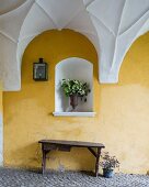 Rustic wooden bench against yellow-painted wall below bouquet in niche in passageway with white vaulted ceiling