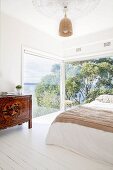 Double bed next to floor-to-ceiling corner windows with panoramic views and ethnic cabinets on white floorboards