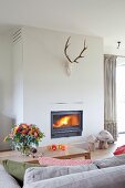 Antlers on wall above lit fire beyond vase of flowers on coffee table