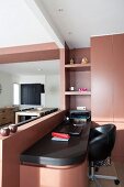 Narrow desk and black swivel chair next to custom fitted cupboards with red-brown fronts; dining area in background