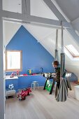 Kind in Hängematte im Kinderzimmer unter dem Dach, mit sichtbarer Holzkonstruktion, im Hintergrund blau getönte Giebelwand
