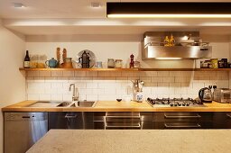 Blick über Theke auf Küchenzeile mit Holzarbeitsplatte vor weiss gefliester Spritzschutzwand, darüber Wandbord aus Holz und Dunstabzug