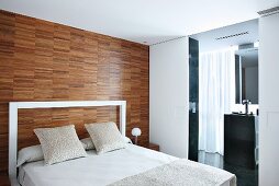 Modern double bed with white, frame-like headboard against wall with parquet-style wood panelling and view into ensuite bathroom