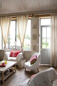 Verschiedene Sessel mit weisser Husse um Couchtisch vor Fenster mit geknoteten Vorhängen in schlichtem Wohnzimmer