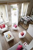 View down from gallery of armchairs with white loose covers, open terrace doors and set table in dining area in open-plan interior