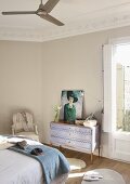 Painting of woman on vintage-style chest of drawers and antique chair at foot of double bed with blanket
