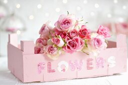 Bouquet of roses in fruit crate painted pastel pink and labelled with decorative lettering spelling 'FLOWERS'