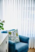 Scatter cushion with large-format floral pattern on light blue armchair in front of white, floor-length curtains on window