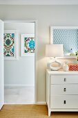 Retro-Tischleuchte auf weisser Kommode neben offener Tür, Blick auf gerahmte Bilder mit dekorativen Ornamentmustern