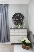 Elegant silver candlesticks in candle lanterns on top of vintage chest of drawers against striped wallpaper and black, vintage-style wall clock in country-house interior