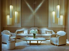 Formal arrangement of armchairs, sofa set, coffee table and groups of pendant lamps in front of geometric wall hanging in white and gold