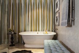 Retro bathtub, side table and upholstered bench in bathroom with striped curtains and marbled wall