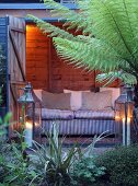 Illuminated wooden shed with sofa, two lanterns and tree fern in garden