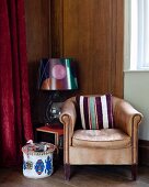 Striped cushion on pale brown leather armchair and table lamp with patterned lampshade on side table in corner of wood-clad room