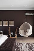 White, wicker, hanging chair in bedroom with wall painted dark brown