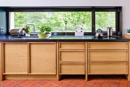 Modern kitchen counter with wooden base units and black worksurface below ribbon window with view of trees