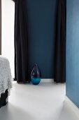Blue glass floor vase on white epoxy resin floor beteen dark blue, floor-length curtains