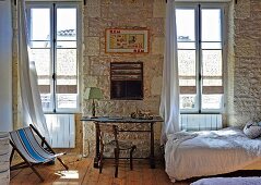 Mediterranean interior with desk and chair in front of rustic stone wall flanked by tall windows; single bed to one side
