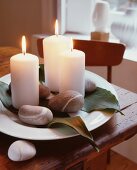 Lit, white candles, pebbles and leaves on ornamental plate