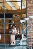 Blick durch Metallgitter auf Frau in Loungebereich einer Loft-Wohnung mit Ziegelwänden und Holzbalkendecke