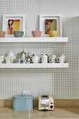 Artistic bowls, framed pictures of women and coffee services on white floating shelves on wall with geometric wallpaper