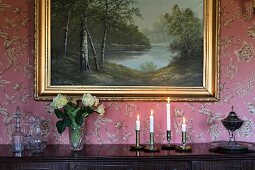 Lit candles and roses on sideboard below gilt-framed landscape painting on wall covered in floral wallpaper