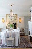 White tablecloth on dining table, white-painted chairs and antique chandelier in traditional dining room