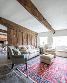 Rustikales Wohnzimmer mit Holzbohlenwand im Hintergrund, davor Sofa und Sessel um Polsterhocker, auf rot-blau gemustertem Teppich