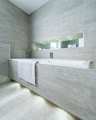 Eingebaute Badewanne mit grauen Fliesen an Frontseite und an Wand, oberhalb schmale, beleuchtete Nische mit Spiegel an Rückwand