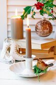 Kerzenhalter aus Porzellan mit Vogelfigur, dahinter Deko-Schlittschuhe, Bücherstapel und Weihnachtskugel an Stechpalmenzweig