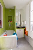 Bathtub next to partition with integrated washstand in bathroom with concrete floor and walls partially tiled in green