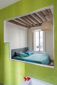 View into simple bedroom with rustic wood-beamed ceiling through opening in green-tiled wall