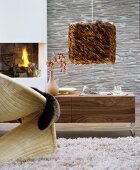 Curved wicker chair on flokati rug, wooden sideboard with extravagantly grained front and pendant lamp with lampshade made from brown feathers in living room